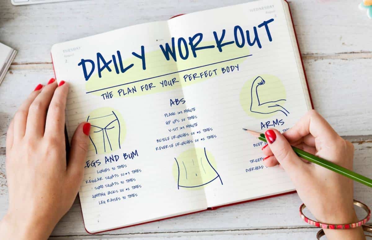Person writing a daily workout plan in a notebook, detailing exercises for abs, arms, legs, and bum.