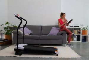A woman in workout gear reads a tablet while sitting on a grey sofa, with a treadmill and houseplants in the background.