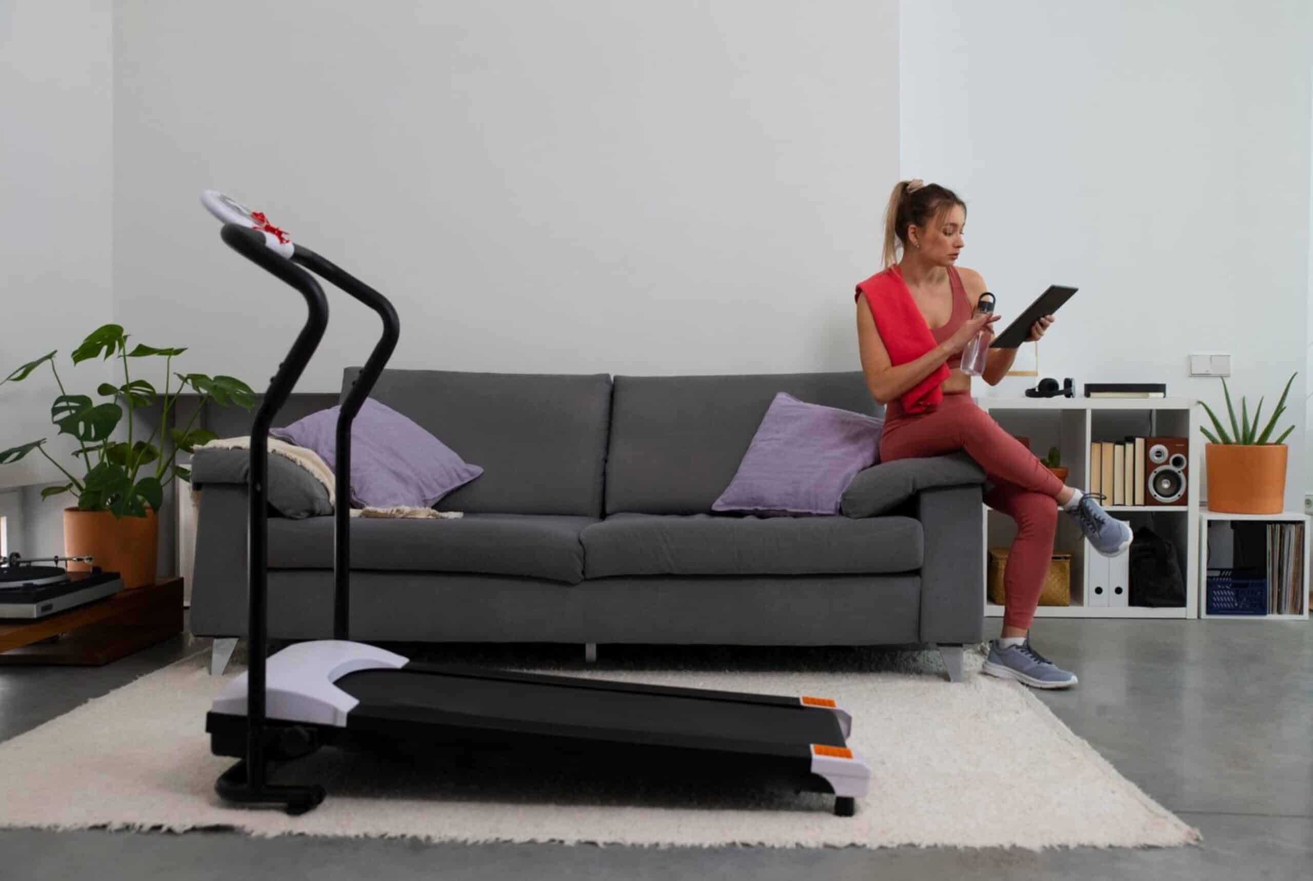 A woman in workout gear reads a tablet while sitting on a grey sofa, with a treadmill and houseplants in the background.