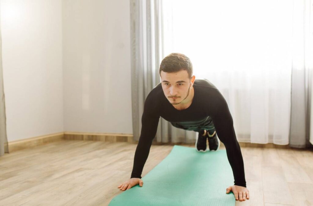 Young man in athletic wear performing a plank exercise on a green yoga mat in a sunlit room.