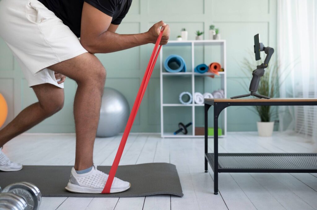Man doing resistance band exercises at home, with exercise equipment and smartphone on tripod in background.