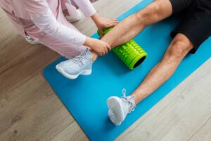 A woman assists a man using a green foam roller on his leg on a blue mat.