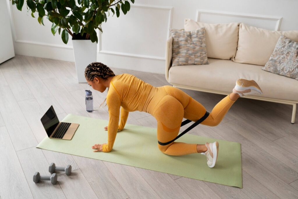 Woman in orange activewear exercising with resistance band at home, following a workout on her laptop.