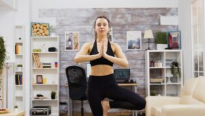 A woman practices yoga in a living room, performing a tree pose with hands in prayer position.