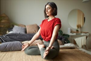 A woman meditates in a cross-legged position on a woven mat in a cozy, plant-decorated bedroom.