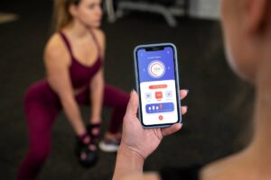 Person using a fitness app on a smartphone in a gym, monitoring exercise progress displayed on screen while another person exercises in background.