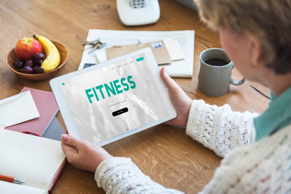 A senior woman holding a tablet displaying a fitness app, with coffee and fruits nearby on a wooden table.