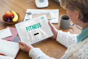 A senior woman holding a tablet displaying a fitness app, with coffee and fruits nearby on a wooden table.