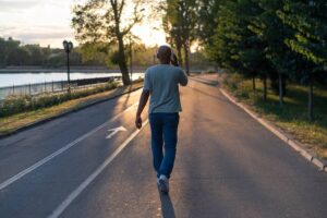 A man walks alone on a tree-lined park path at sunset, wearing headphones and casual clothing.