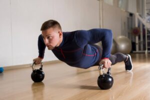 Man in blue sportswear does push-ups with kettlebells in a gym.