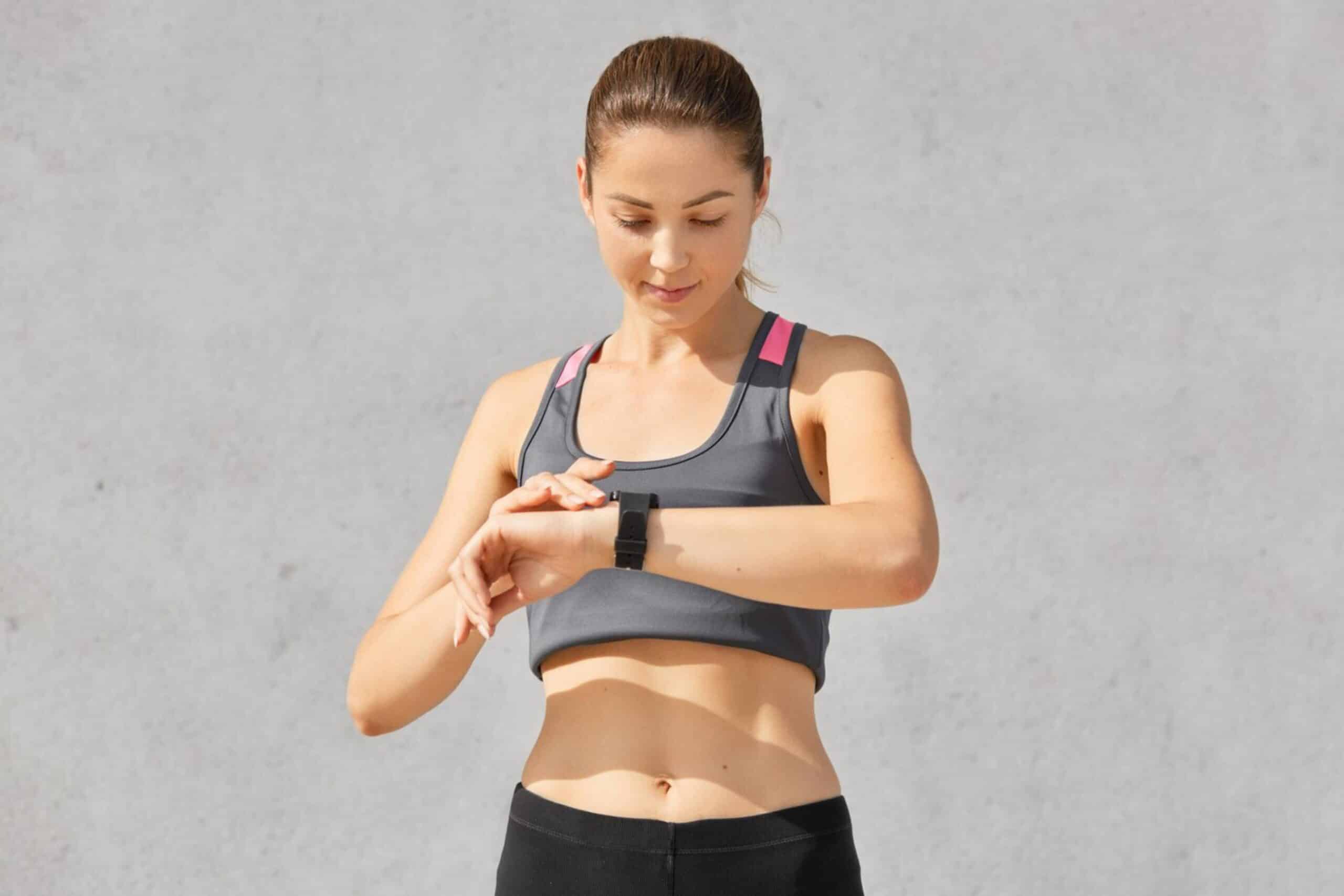 Young woman in sports attire checking fitness tracker against a plain background.