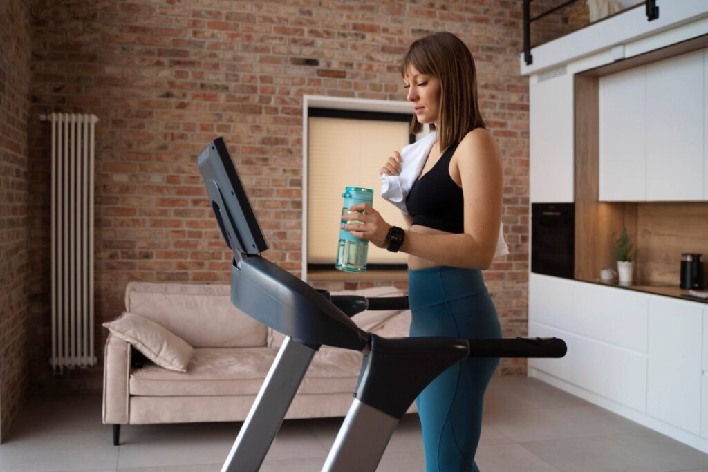 Woman holding a water bottle while standing on a treadmill in a modern home with brick walls.