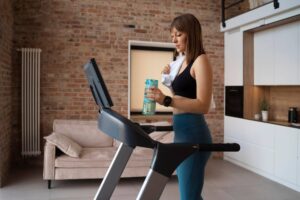 Woman holding a water bottle while standing on a treadmill in a modern home with brick walls.