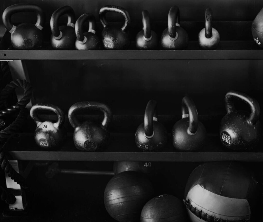 A black and white photo showing rows of various sized kettlebells and exercise balls on shelves at a gym.