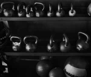A black and white photo showing rows of various sized kettlebells and exercise balls on shelves at a gym.