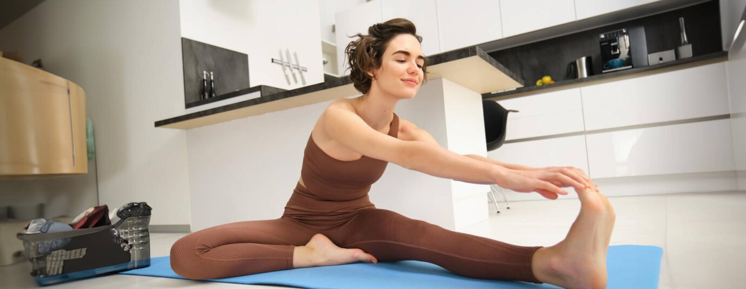A woman in brown activewear stretches on a blue mat in a modern kitchen. 