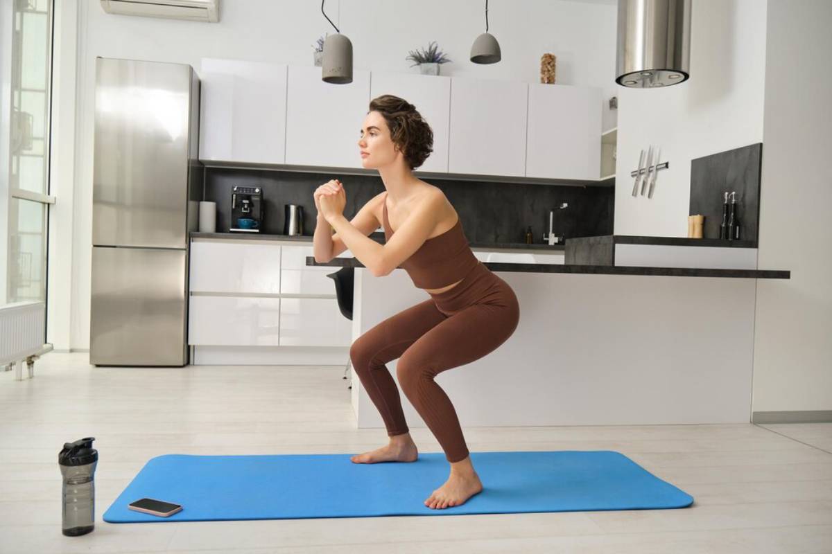 A woman in brown sportswear performs squats on a blue mat in a modern kitchen.