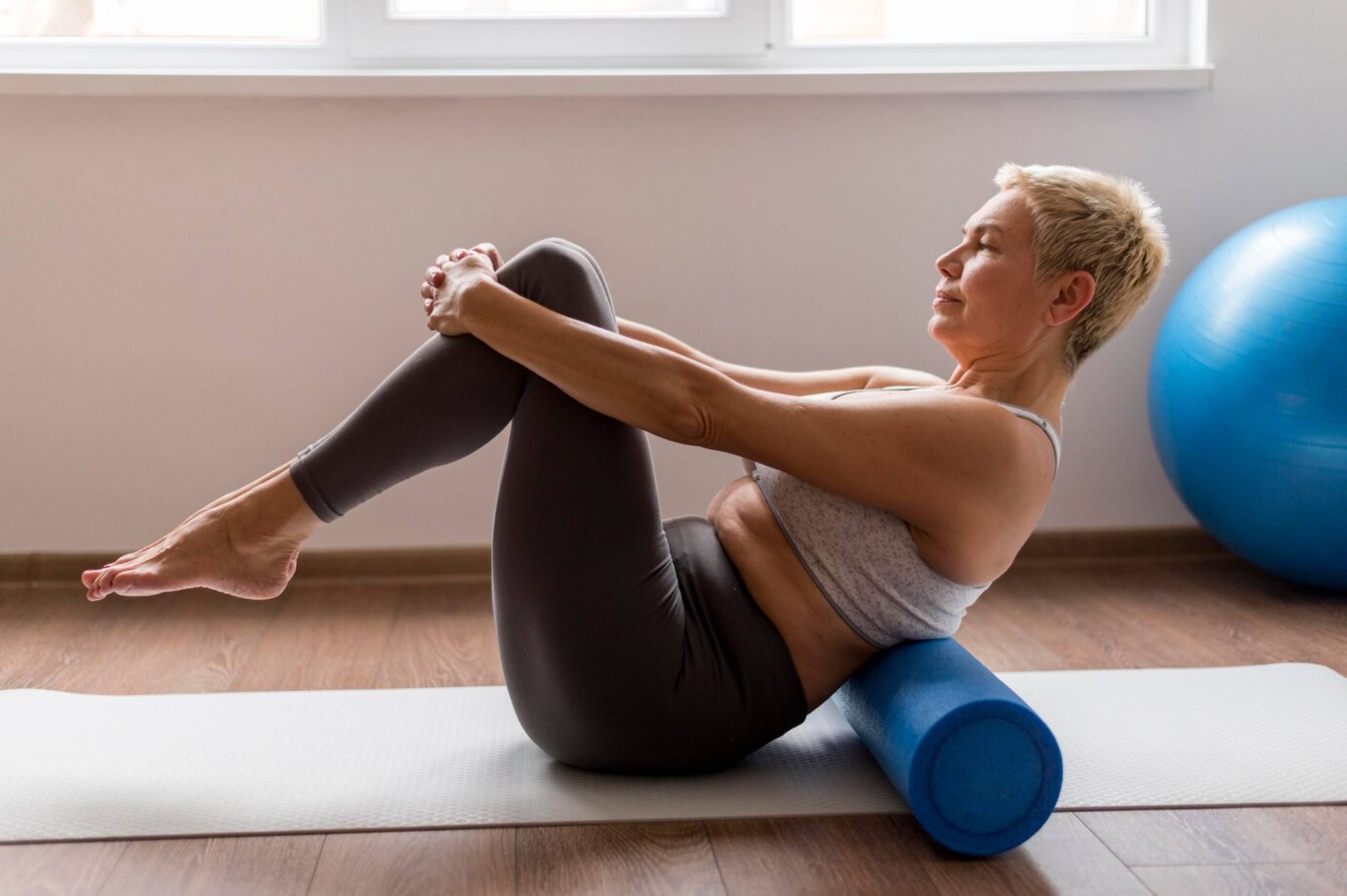 Mature woman performs pilates exercise on foam roller in a bright room.