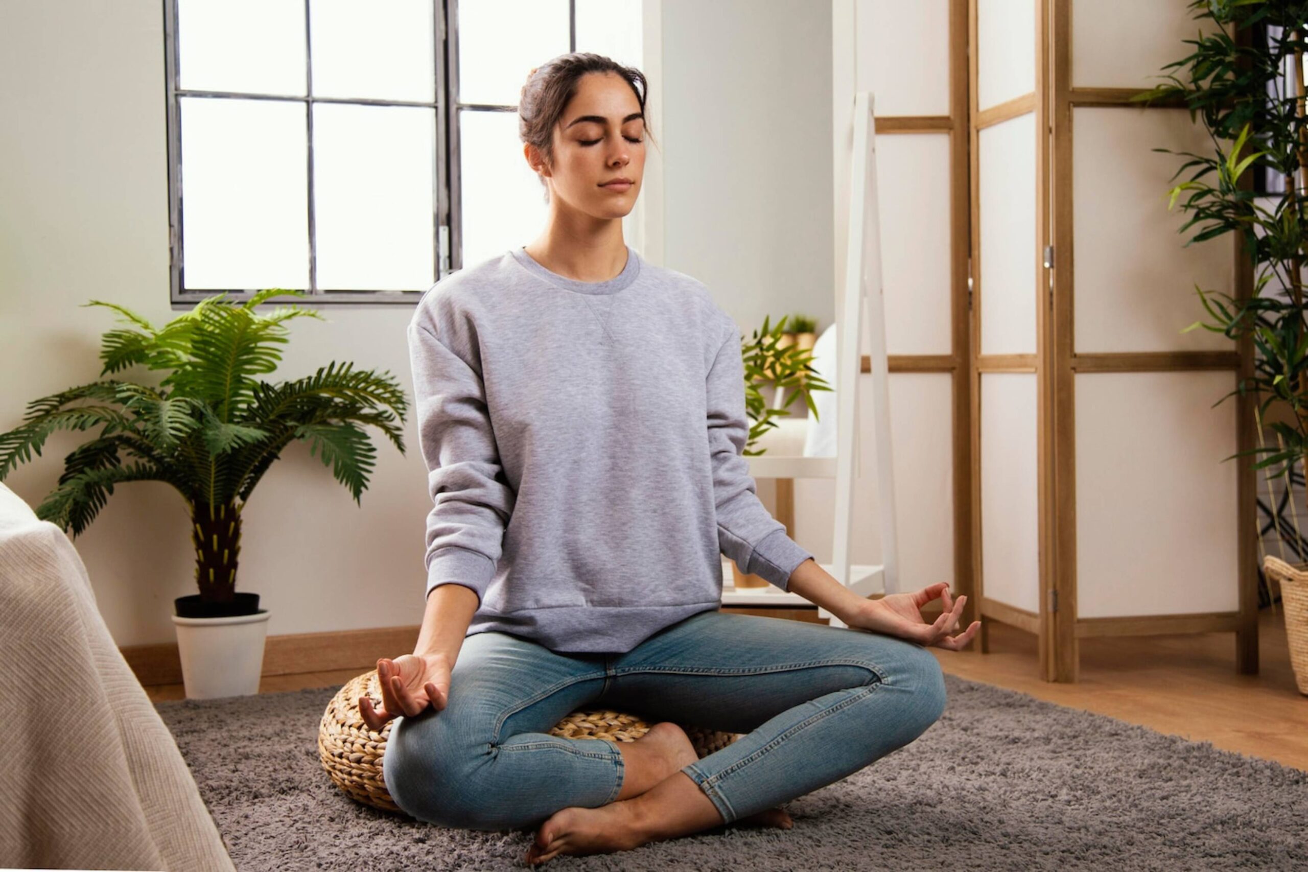  Woman meditating in a bright room with plants and a wicker mat.