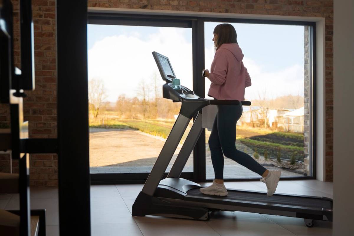 Woman in pink hoodie and leggings walking on a treadmill at home, facing a large window overlooking a sunny rural landscape.