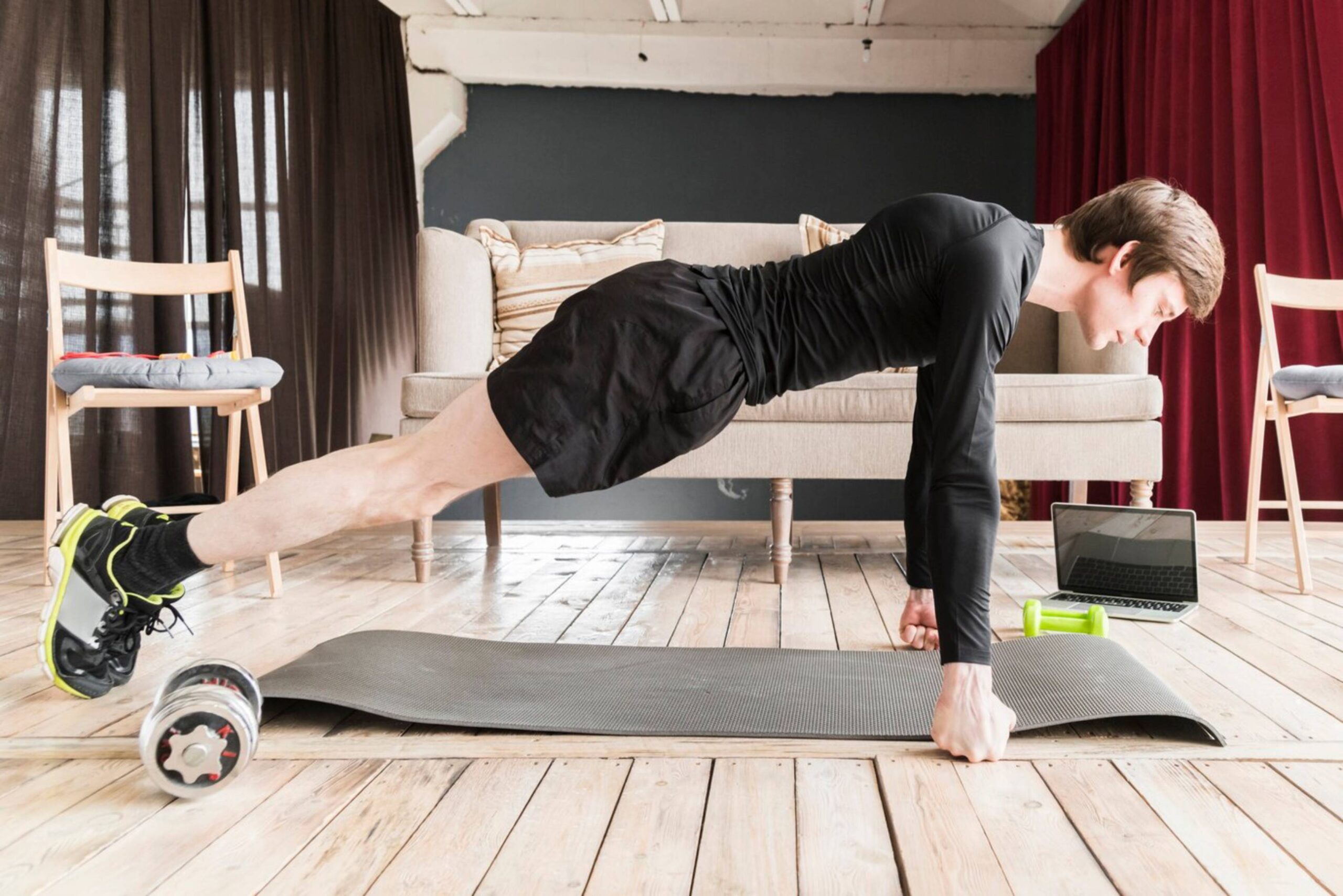  Man in black sportswear performing a plank exercise at home with a laptop and exercise equipment nearby.