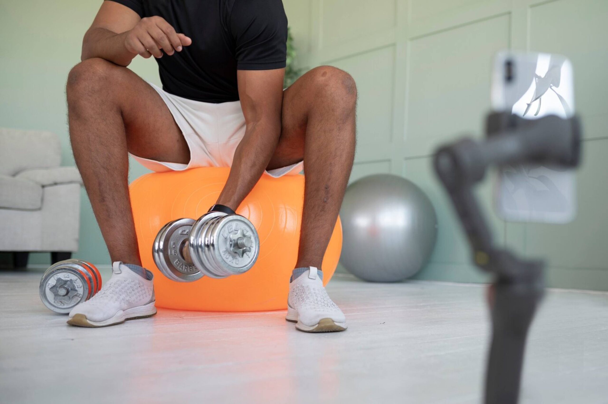  Man recording fitness video, lifting dumbbell while sitting on an exercise ball, smartphone on tripod in foreground.