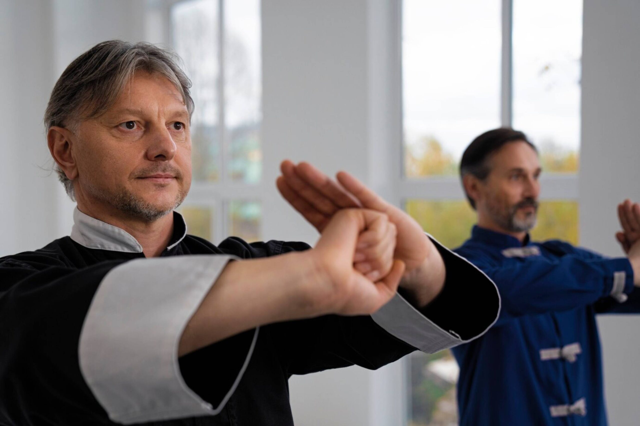 Two men practicing martial arts in a brightly lit room, performing hand forms in traditional outfits.