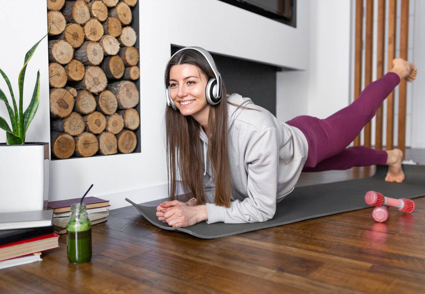  Woman in headphones smiling while doing an exercise on a yoga mat at home, with dumbbells and a green smoothie nearby.