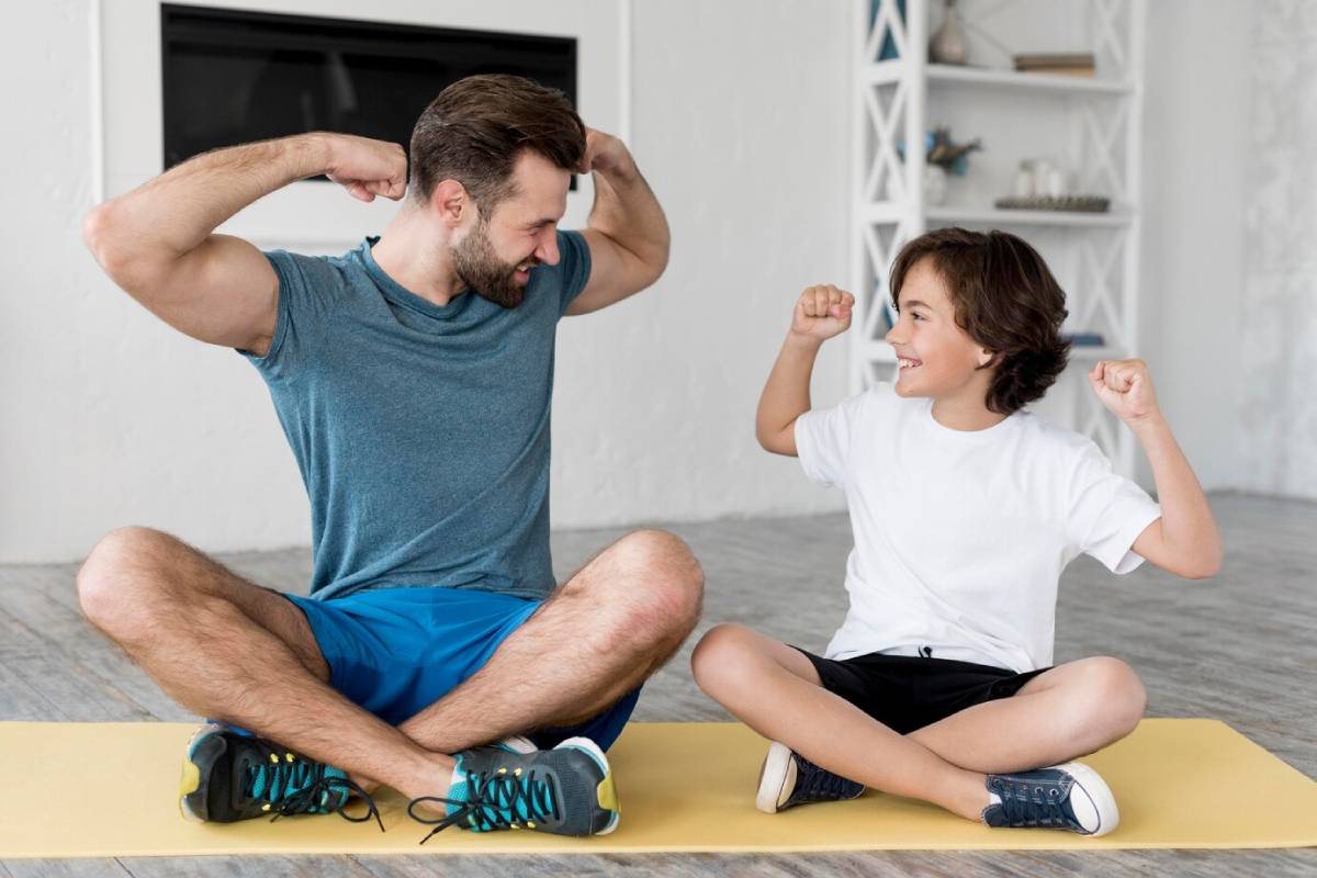 A man and boy sitting on a yoga mat, flexing their muscles and smiling at each other. 