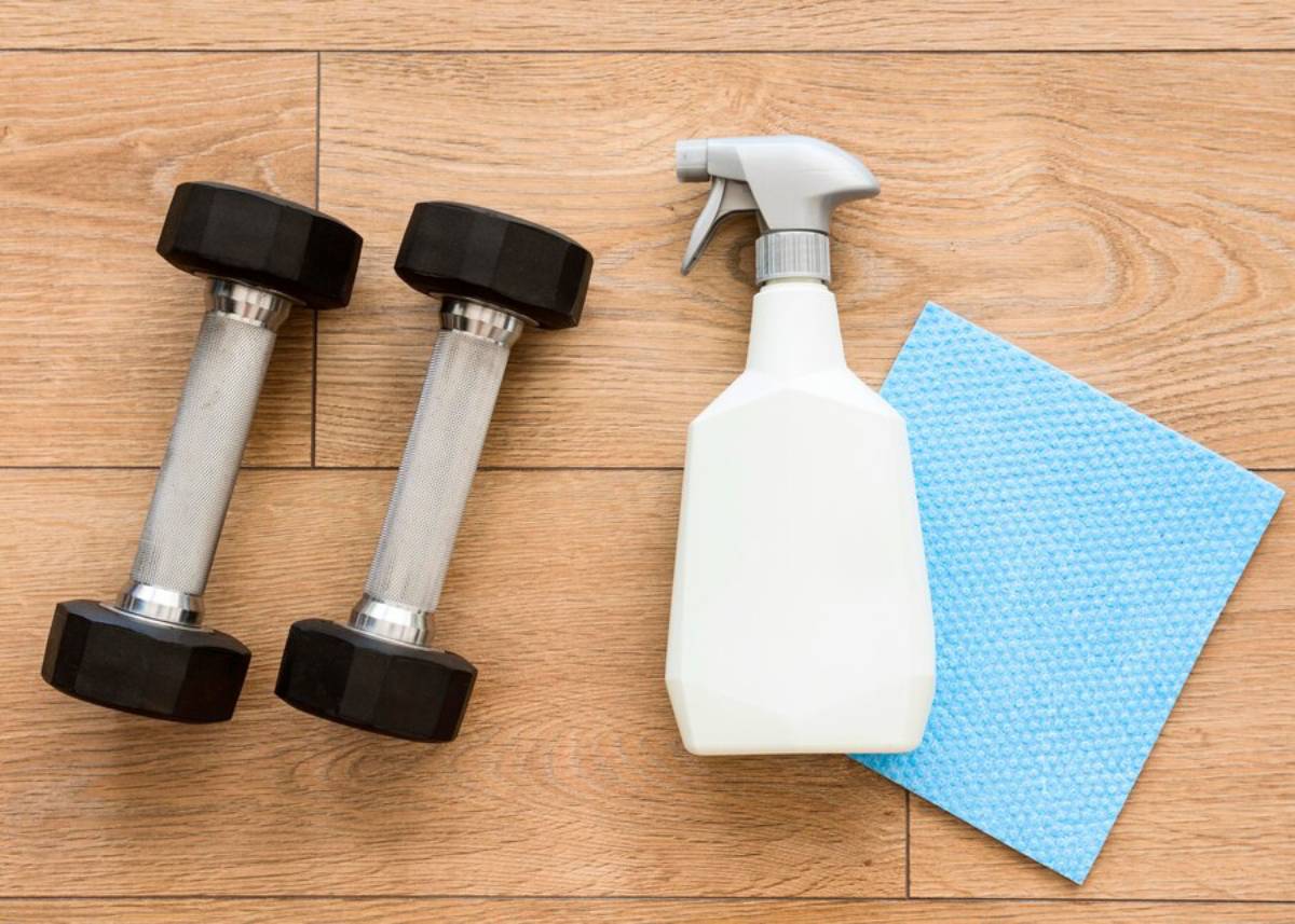 Two dumbbells beside a spray bottle and a blue cleaning cloth on a wooden floor.