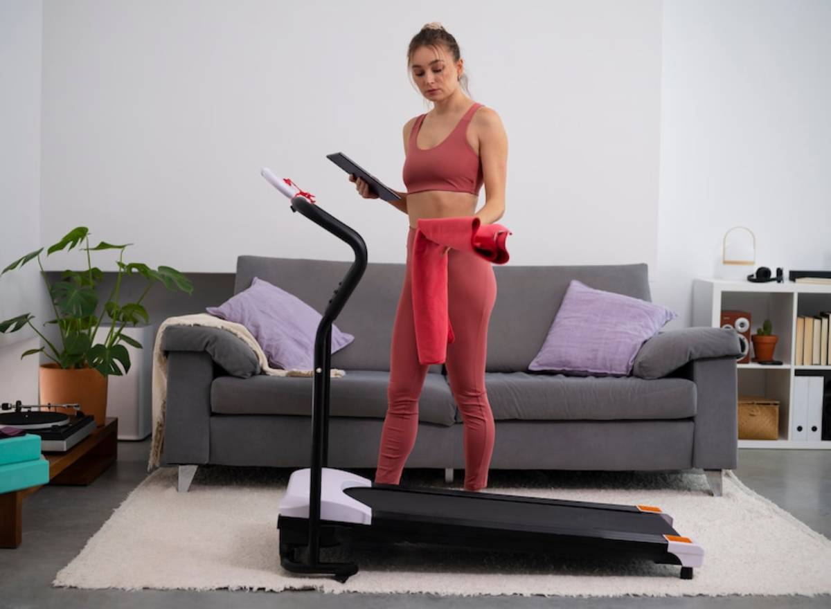 Young woman in activewear checking a tablet while standing on a treadmill in a modern living room.