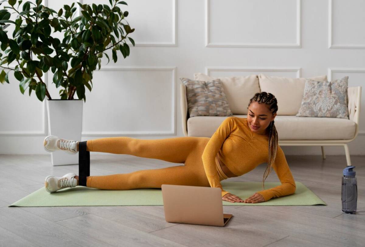 Woman in yellow sportswear stretching on a mat while looking at a laptop in a bright living room. 