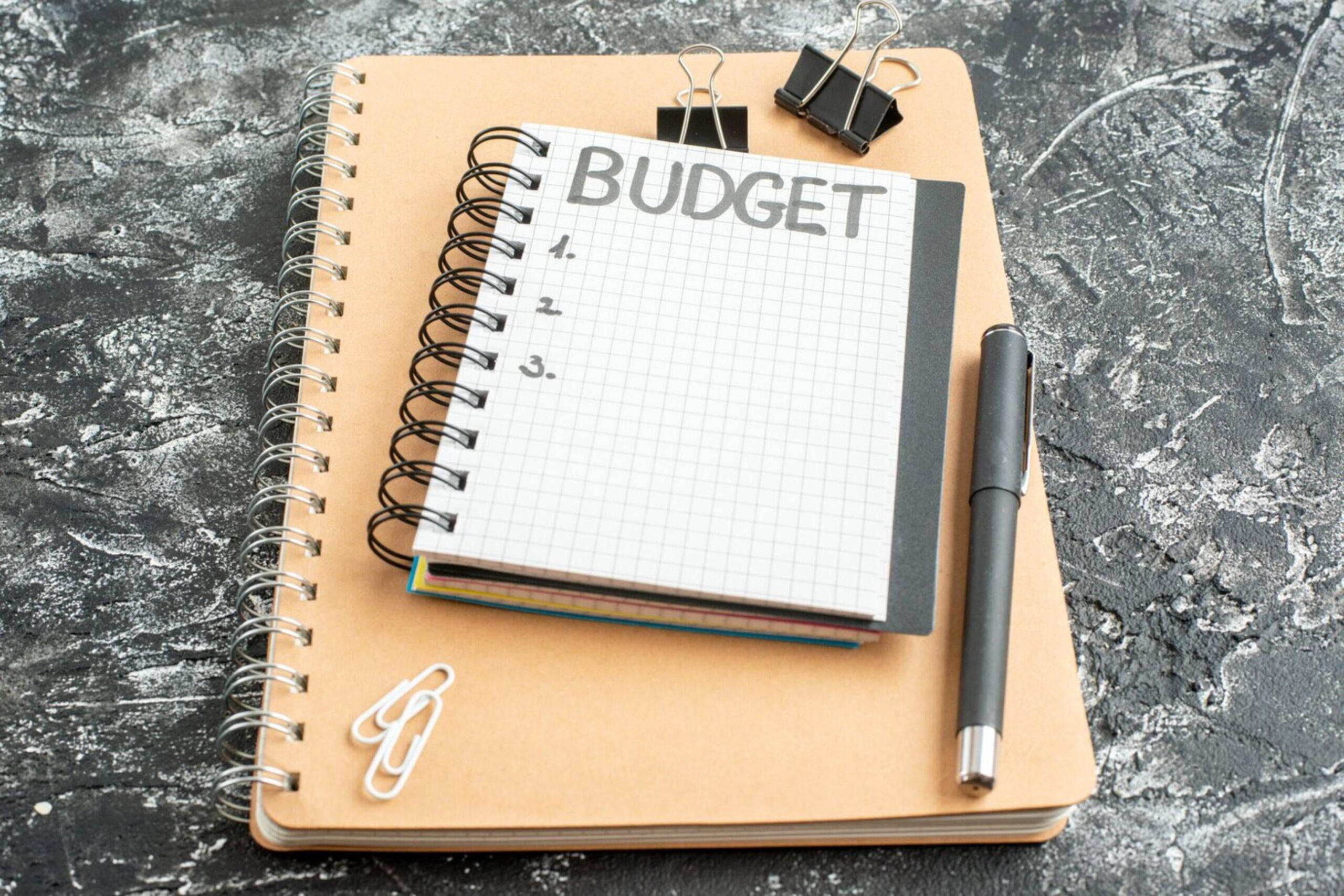  Notebook with  budget title on textured table, pen, paper clips, and binder clips.