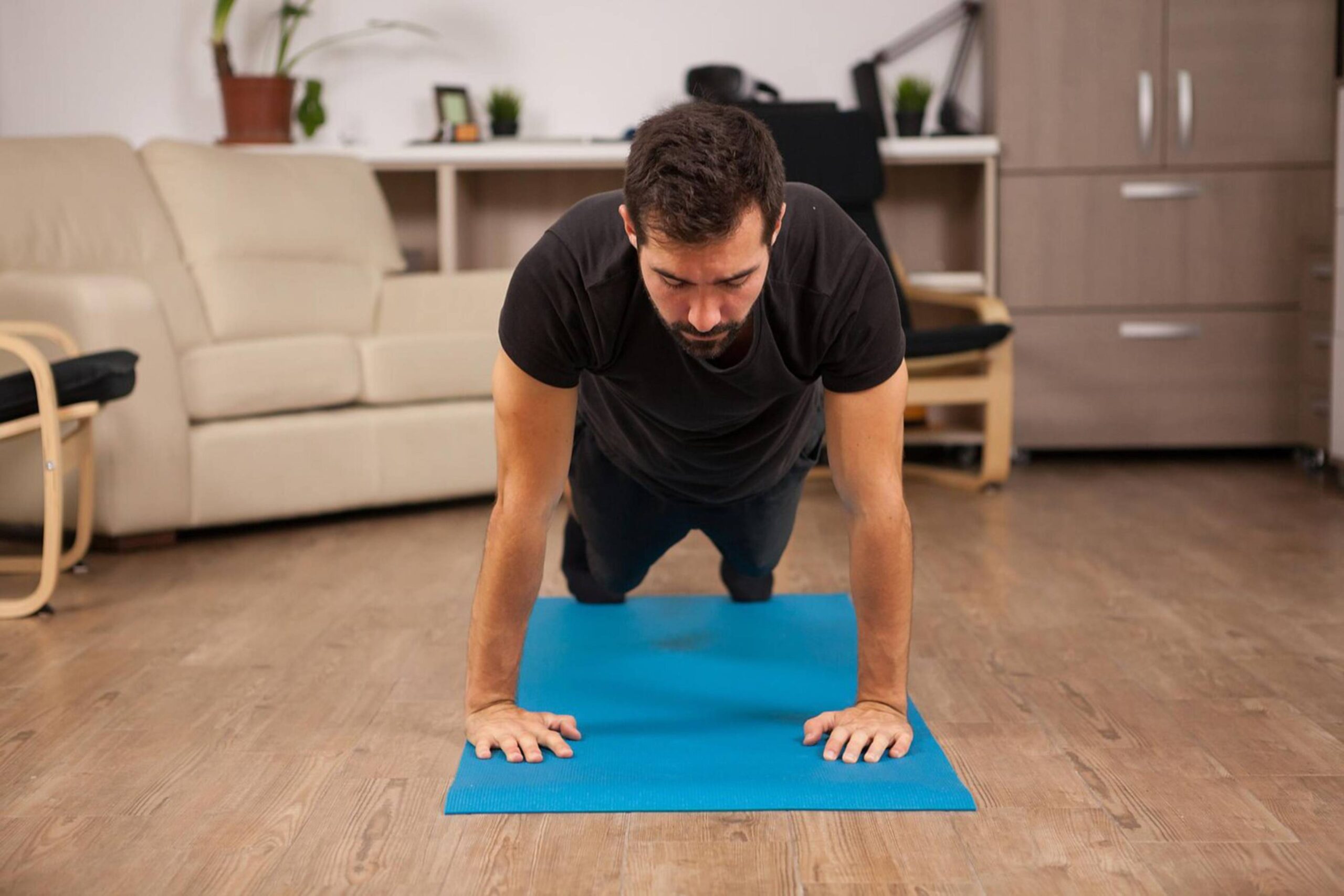 Man in black t-shirt doing push-ups on a blue mat in a home living room. 
