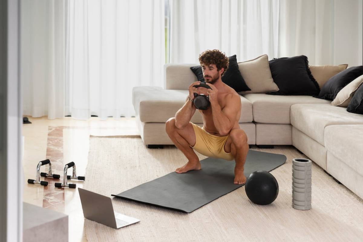  Man doing a kettlebell workout at home, with exercise equipment and a laptop on a mat.