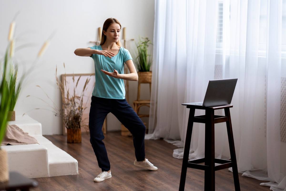 Woman practising tai chi in a peaceful home environment, following a tutorial on a laptop.