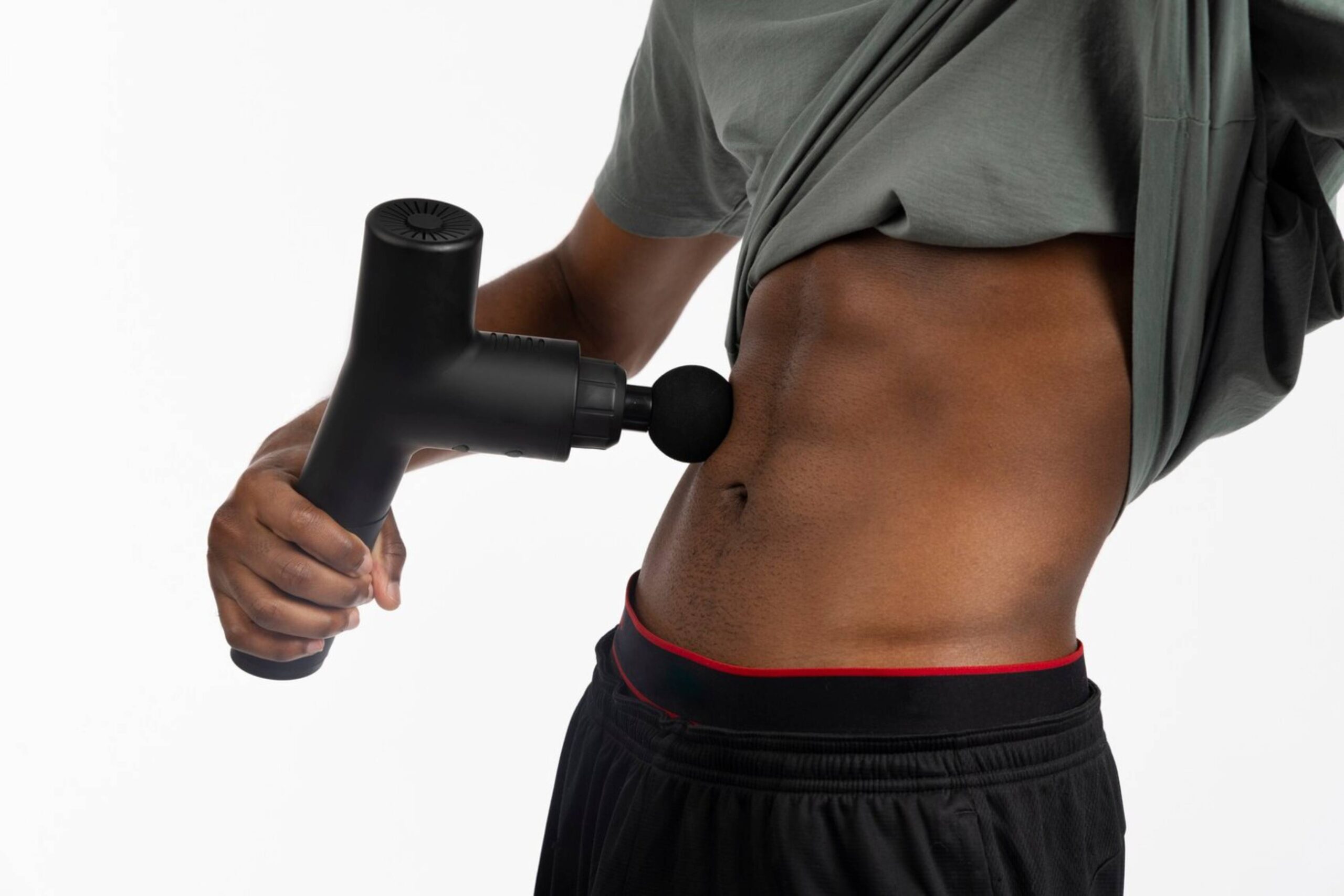  Man using a massage gun on his torso, lifting his shirt to reveal his abs.