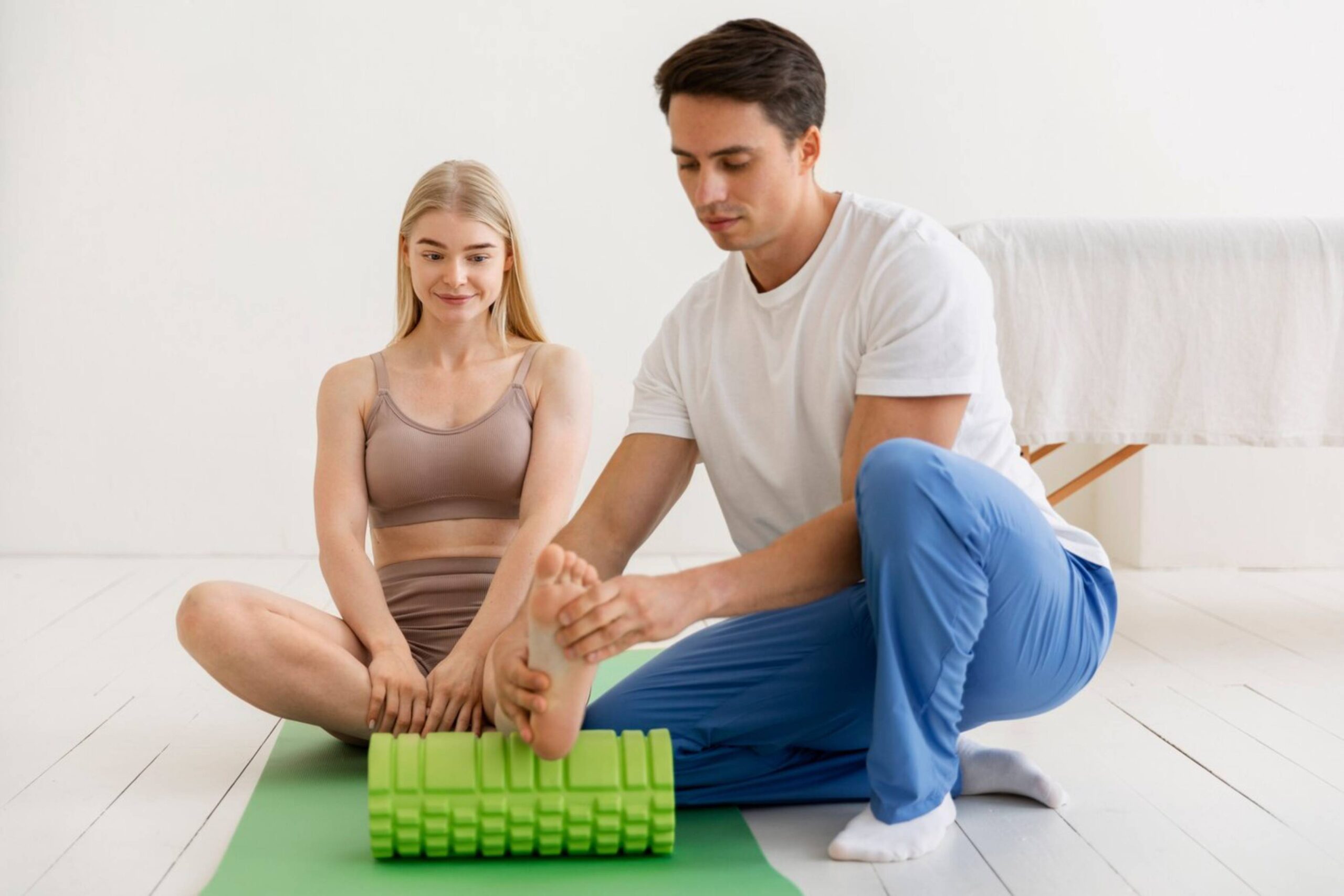  A man assists a woman with a foot stretch on a foam roller in a bright room.