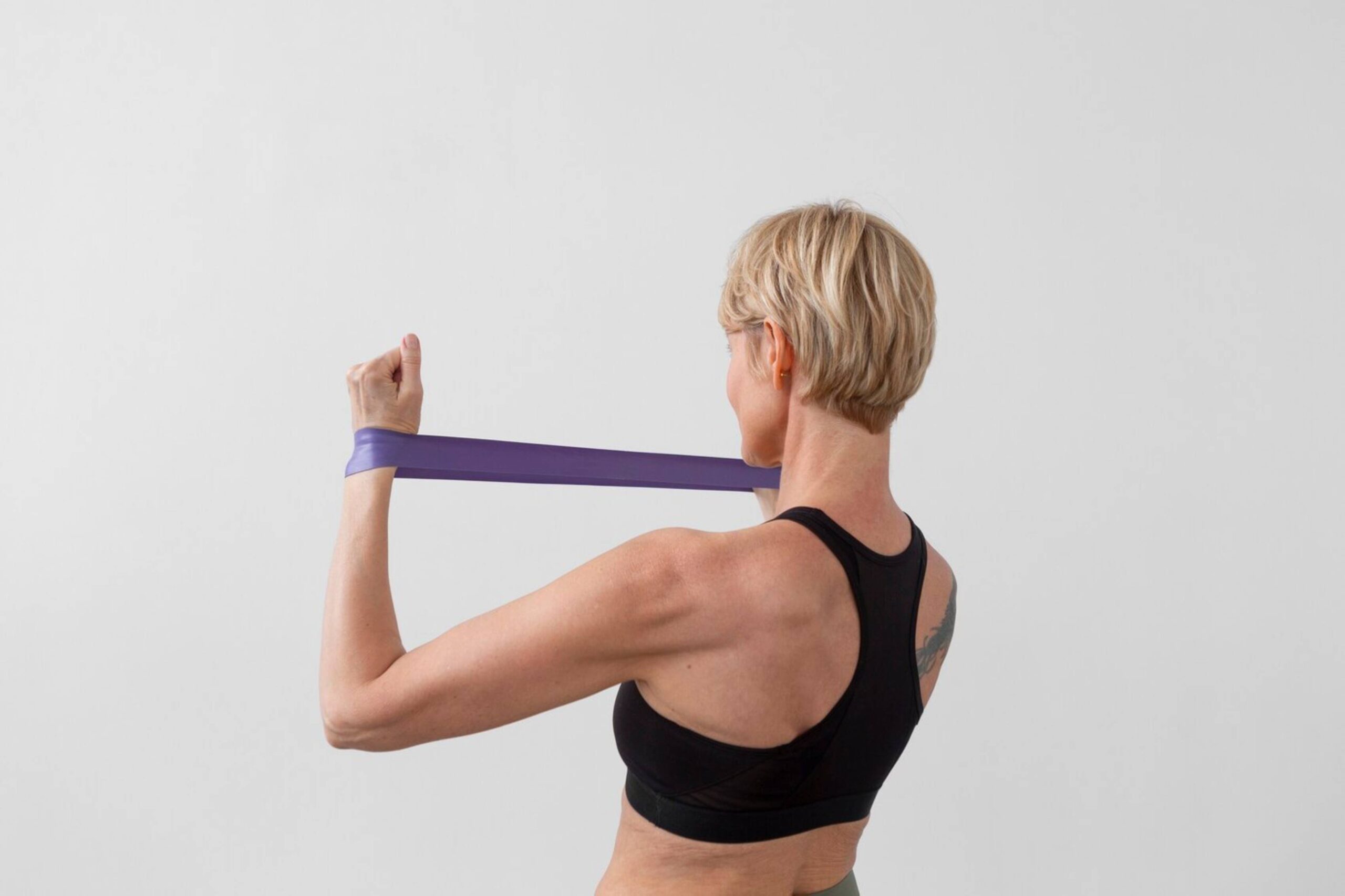 A woman with short hair exercises with a purple resistance band against a plain background. 