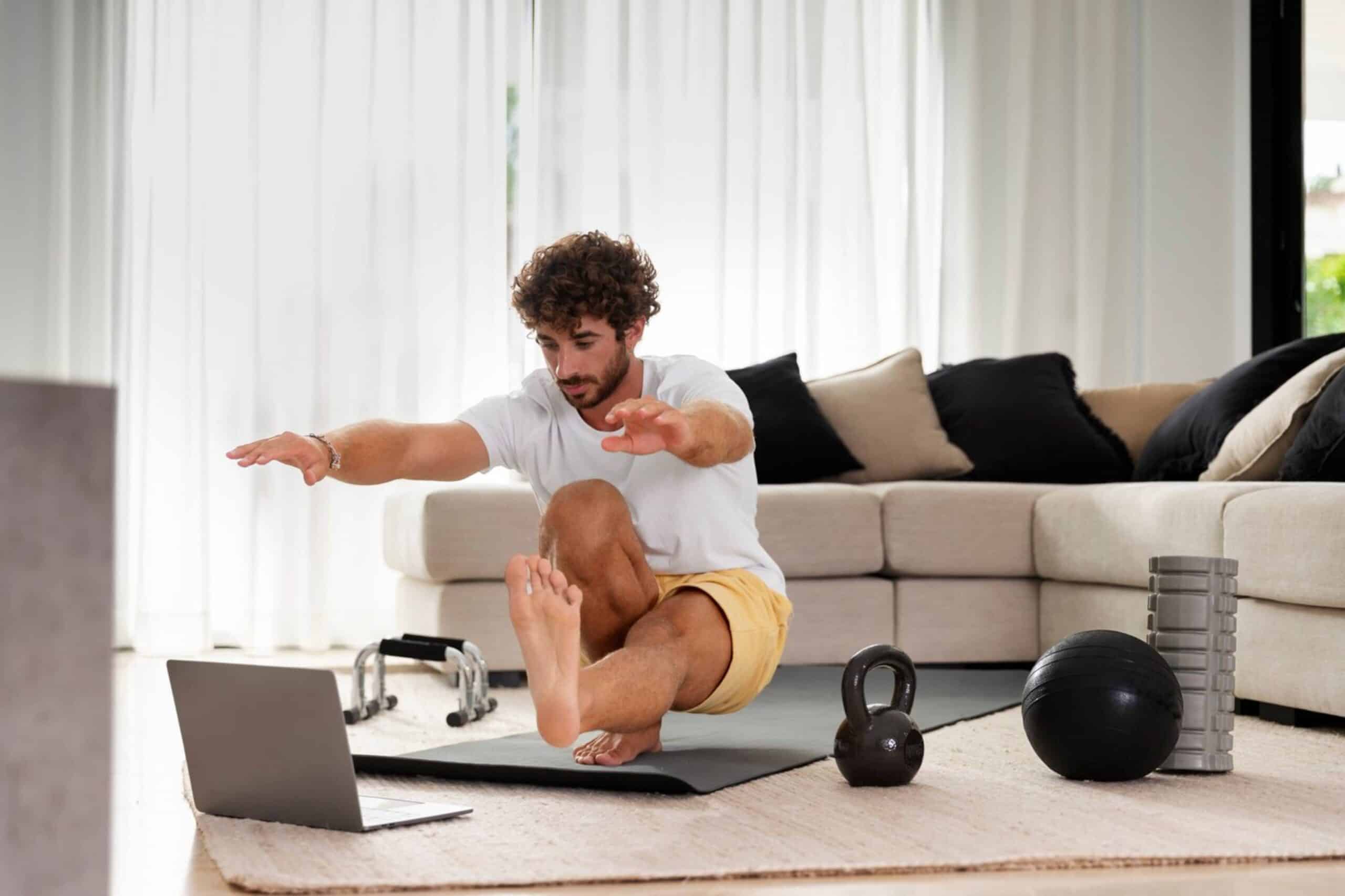 Man doing yoga at home while watching a tutorial on a laptop, with fitness equipment nearby.
