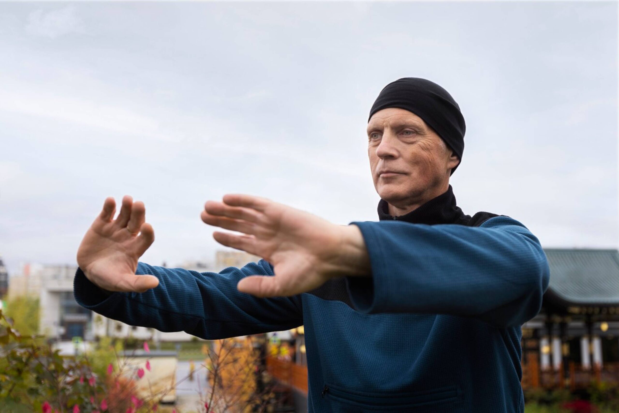  Older man practicing tai chi outdoors, extending his arms with focused expression, wearing a blue top and black headband.