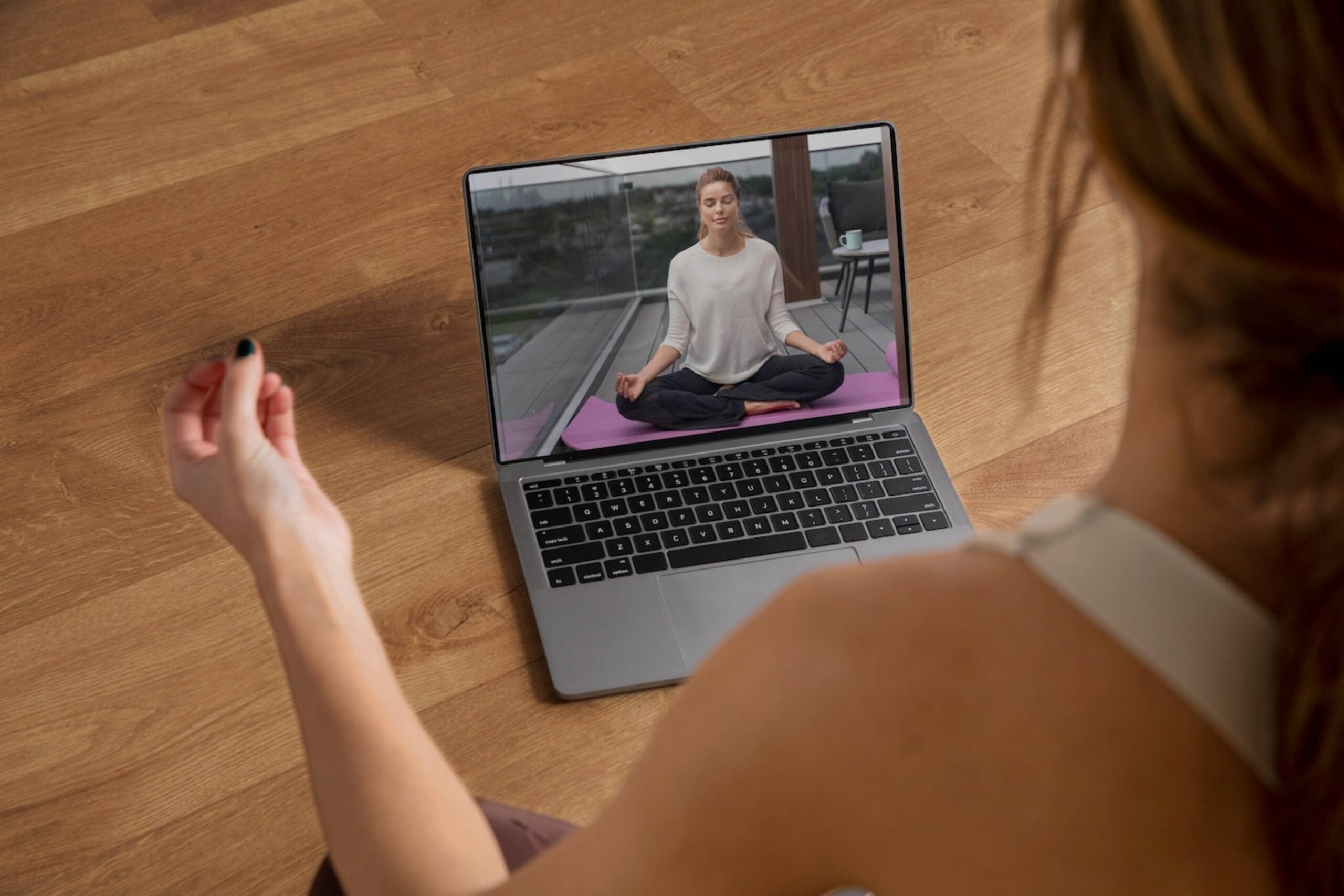 Woman watches an online yoga class on a laptop, imitating instructor's meditation pose at home. 