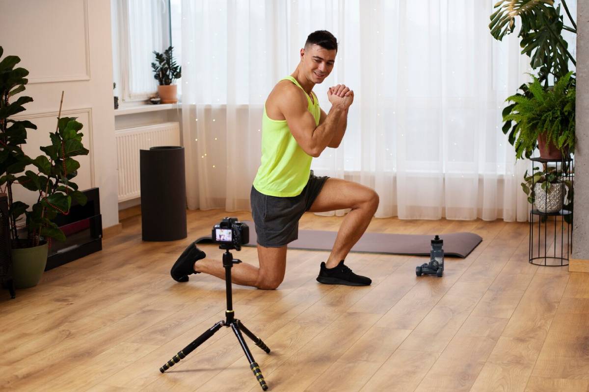 Man in a lime tank top demonstrating a lunge in a home workout session, recording with a camera on a tripod.