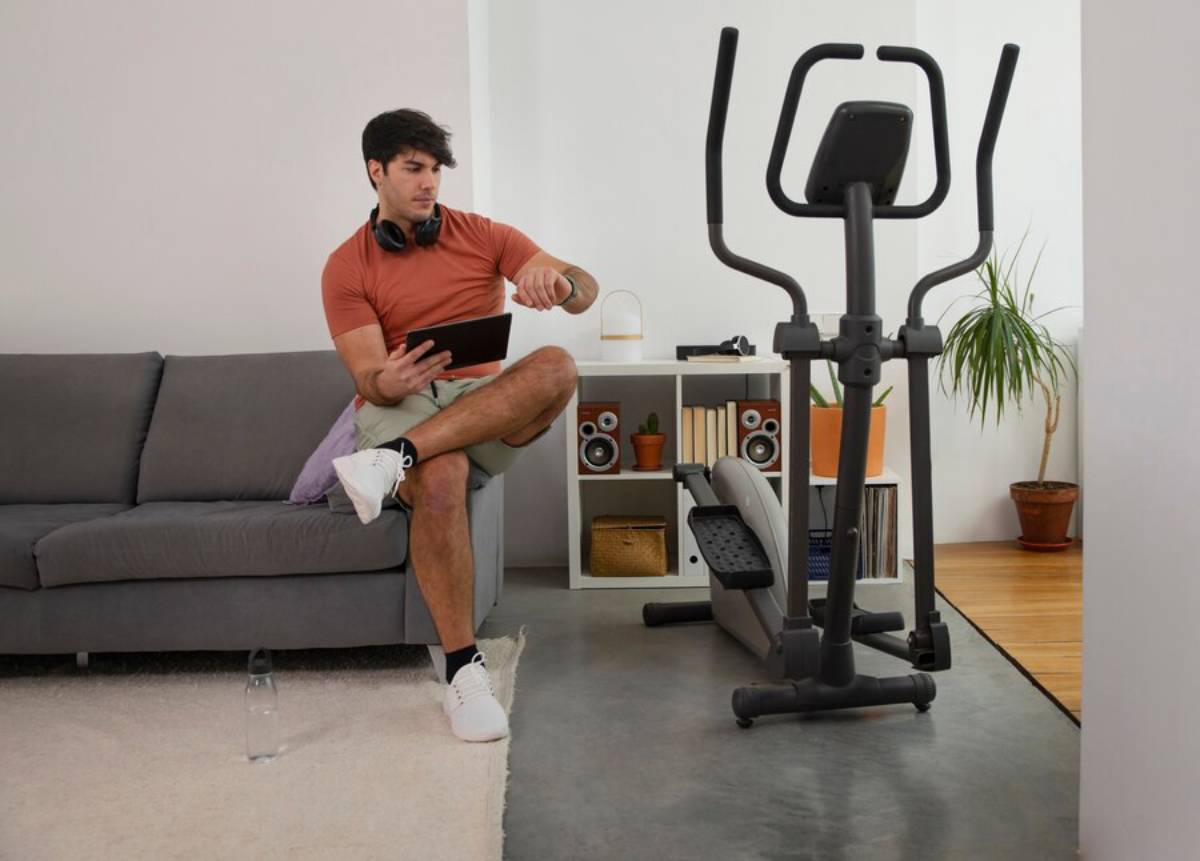  Man sitting on a couch using a tablet with an elliptical machine nearby in a home setting.