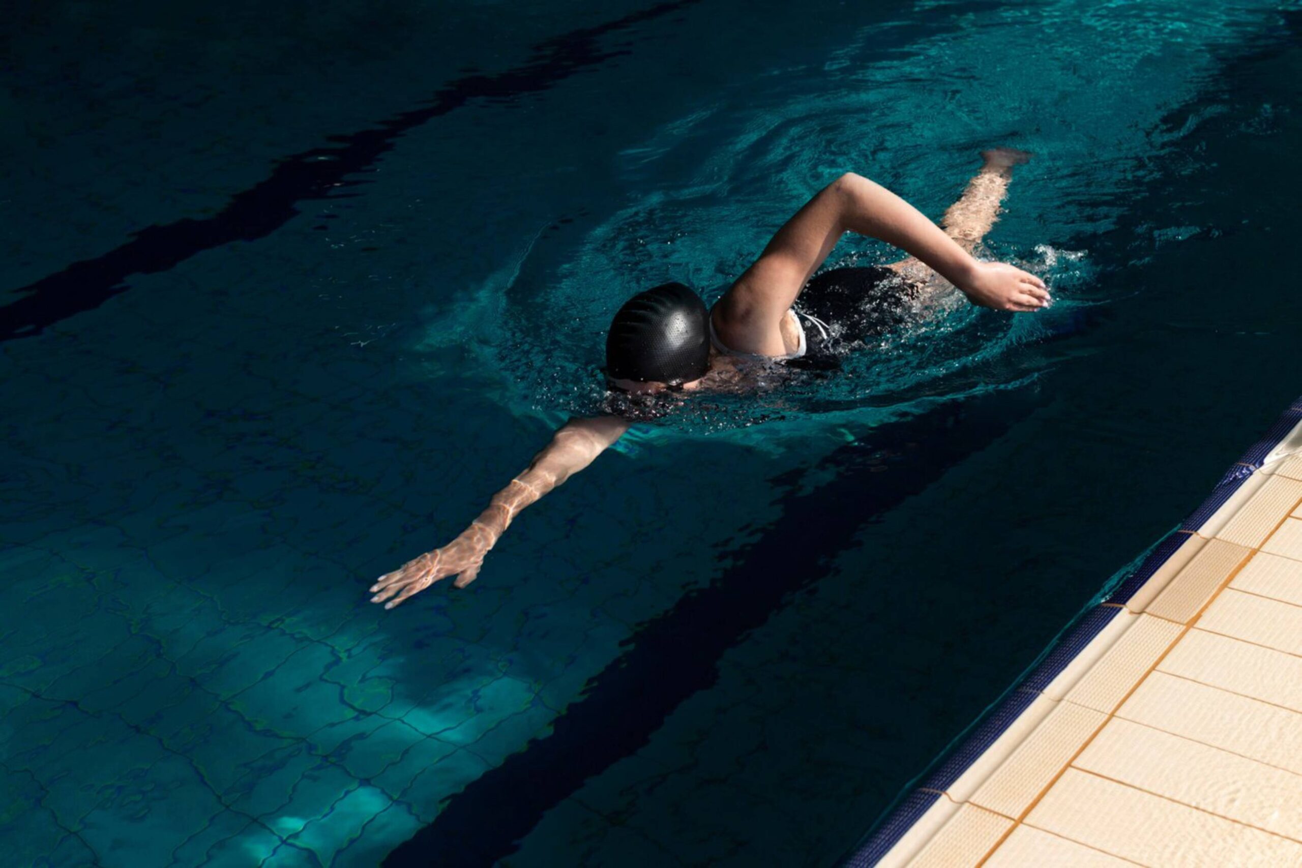 Person swimming in a sunlit outdoor pool, wearing a black swim cap.