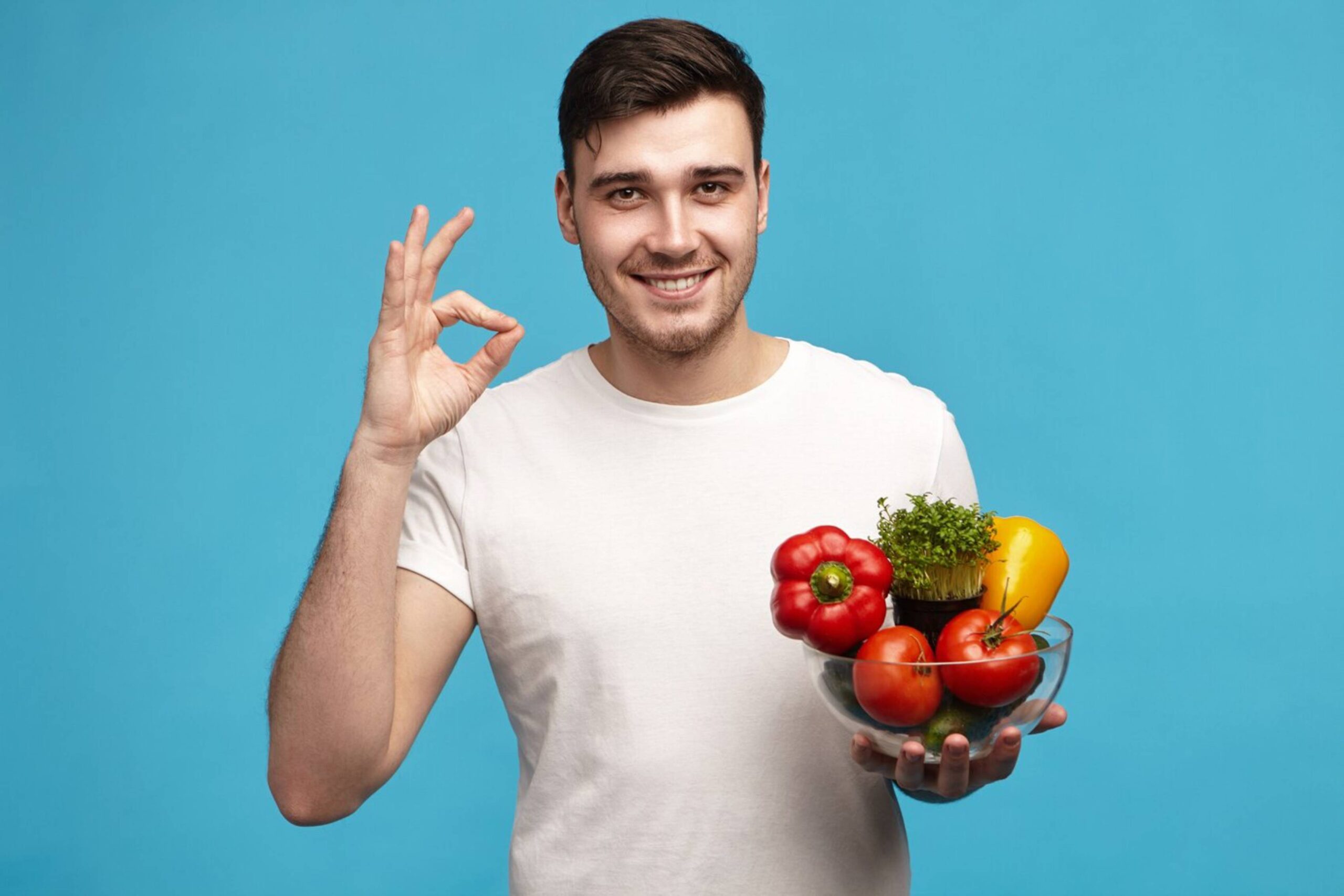 Young man holding a bowl of fresh vegetables and making an OK gesture against a blue background. 