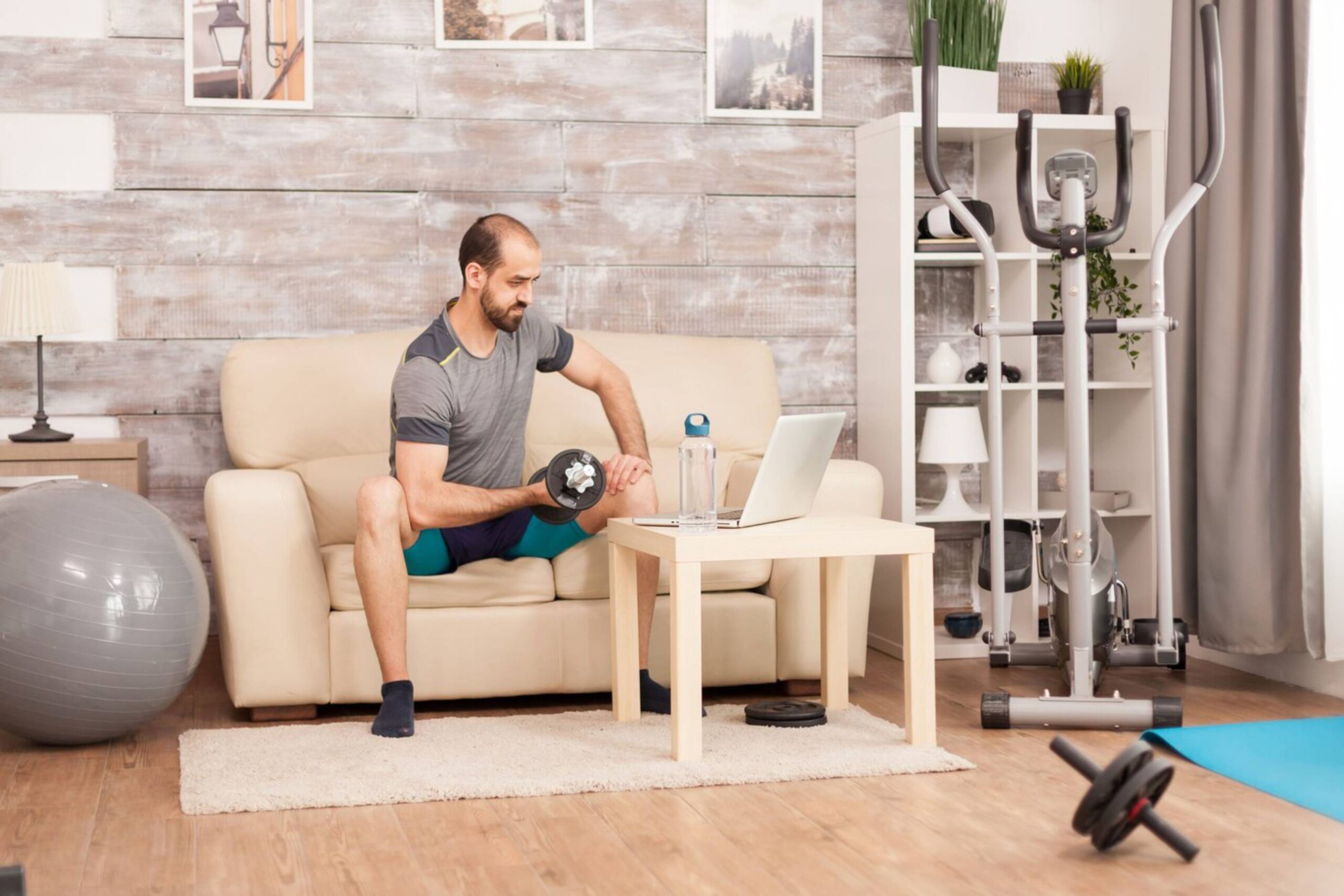 Man exercising with dumbbell while looking at laptop in a home living room setup.