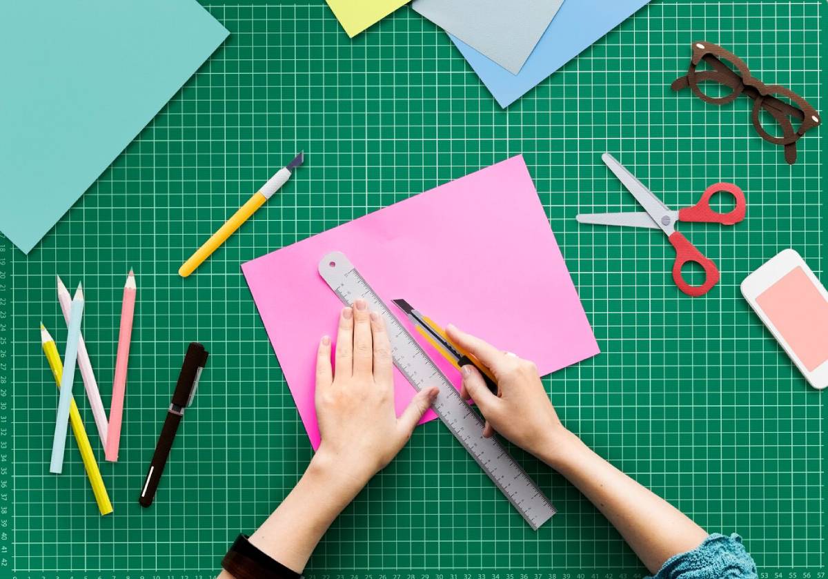A person using scissors to cut paper, surrounded by various crafting supplies on a table.
 